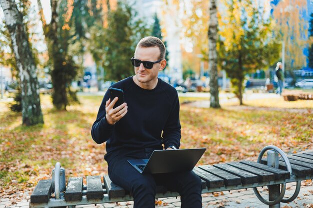 Attractive young man in a black sweater and sunglasses he is sitting in an autumn park on a bench and works on a laptop