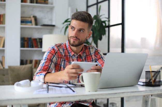 Attractive young man sitting at desk at home office and using mobile phone for cheking social nets.