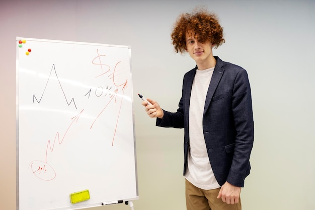 Attractive young man standing in meeting room holding marker and pointing to graph drawn on flip chart Confident businessman