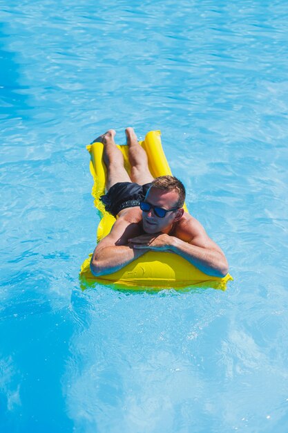 Attractive young man in sunglasses and shorts is relaxing on an inflatable yellow mattress in the pool Summer vacation