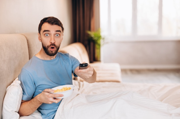 Attractive young man with beard eating breakfast in bed and look disappointed shocked whilst watching news