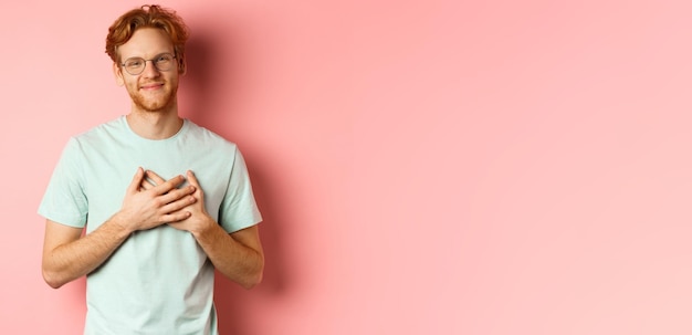 Attractive young man with ginger hair holding hands on heart and smiling grateful saying thank you e