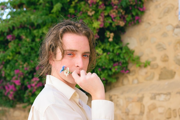 Attractive young man with makeup poses with flowers looking at camera