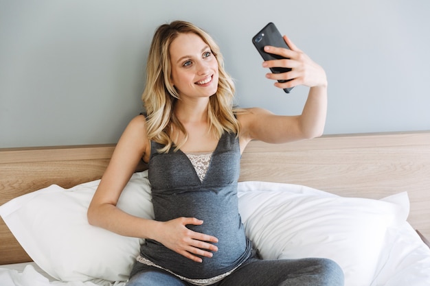 Attractive young pregnant blonde woman relaxing on bed at home, taking a selfie