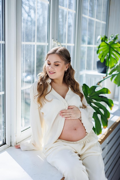 An attractive young pregnant woman in a light white suit is sitting on the windowsill Beautiful pregnant woman stroking her belly