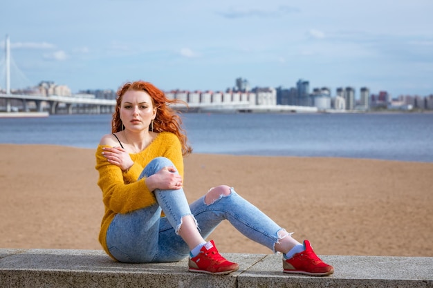 Attractive young redhead woman sitting outside on waterfront wearing yellow