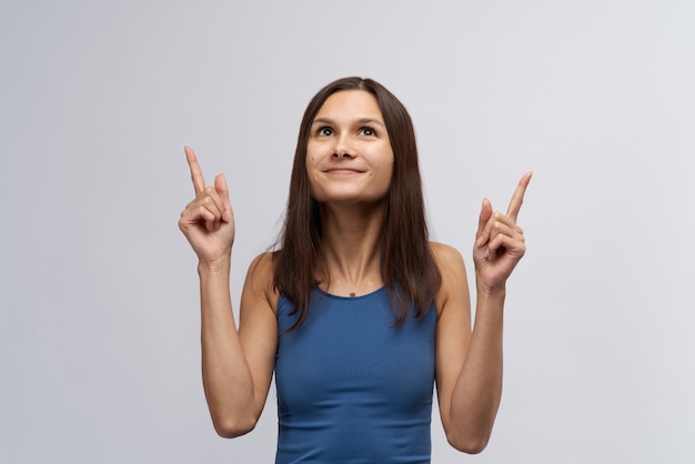 Attractive young smiling woman girl in tight blue t-shirt and with long straight dark hair looks up and forefinger pointed upward. The concept of ad space and the view direction