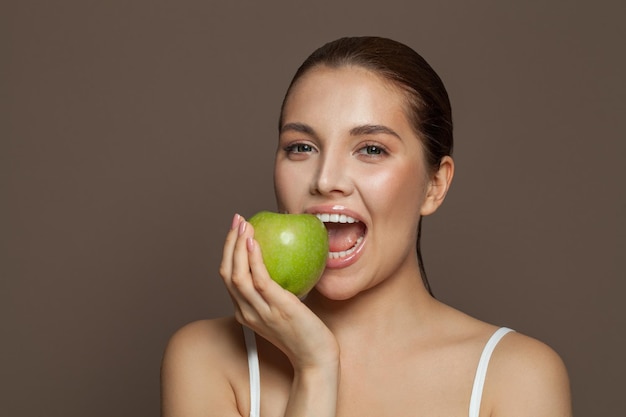 Photo attractive young woman biting green apple and smiling on brown background