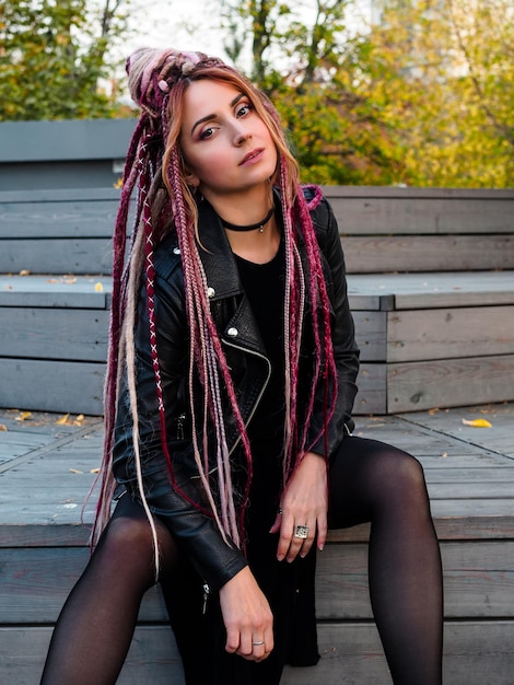 Attractive young woman in black clothes with pink de-dreadlocks sits on wooden steps and looks at the camera