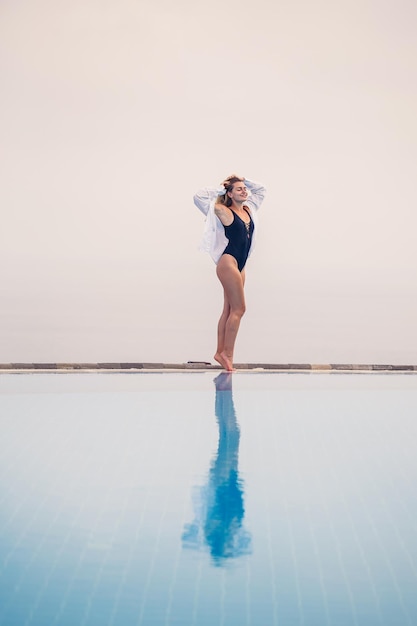 An attractive young woman in a black swimsuit and white shirt stands near the pool with a panoramic view Selective focus