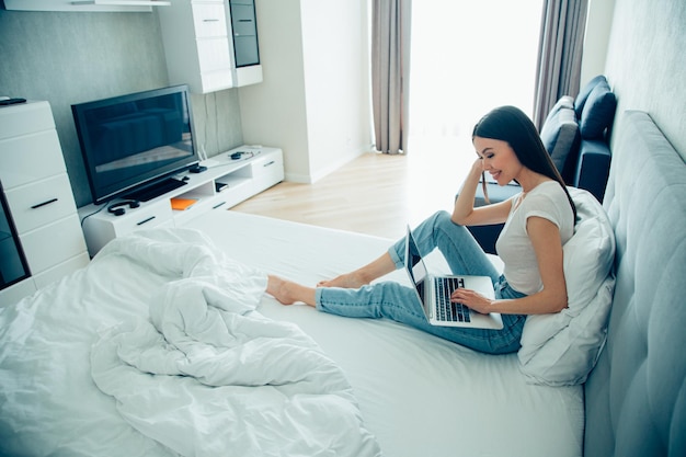 Attractive young woman in casual clothes sitting on a bed at home and having a modern laptop on her lap