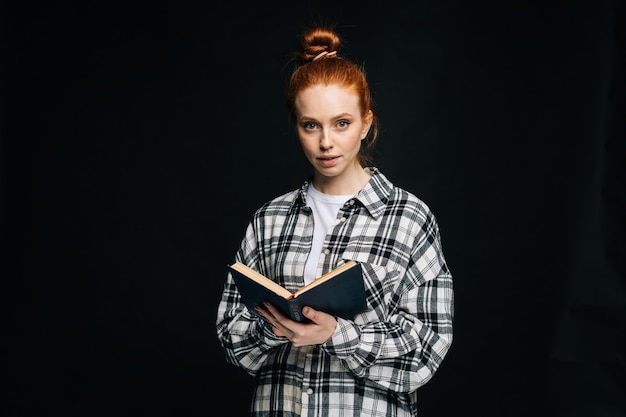 Attractive young woman college student holding opened book and looking at camera on black background