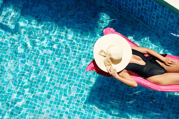 Attractive young woman covering face with straw hat when sunbathing on floating mattress in swimming pool