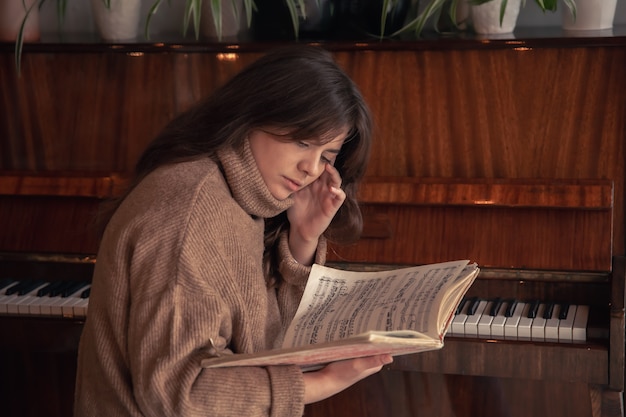 An attractive young woman in a cozy sweater sits near the piano and looks at the notes.