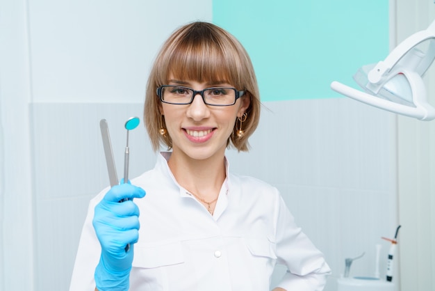 Attractive young woman in dental clinic with a tool in hand in glasses smiling caucasian woman worke...