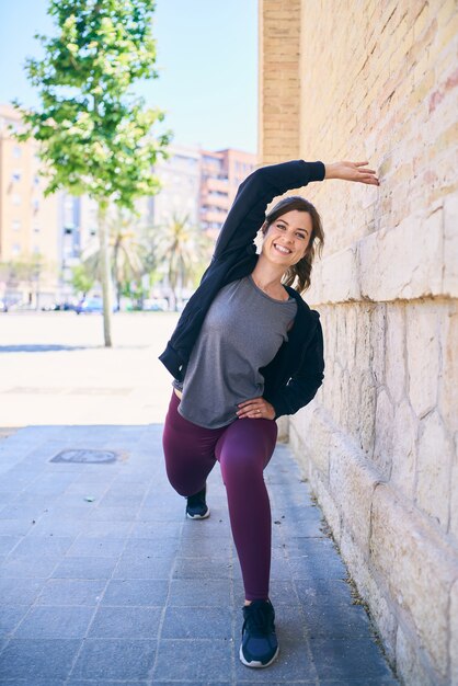 Attractive young woman doing stretching workout in front of bricked wall. Urban location. Fitness model exercising, sportswear and black hoodie. Leg and arm stretching.