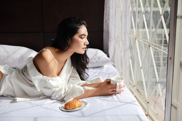 Attractive young woman eating coffee and croissant for breakfast in bed at home in the morning