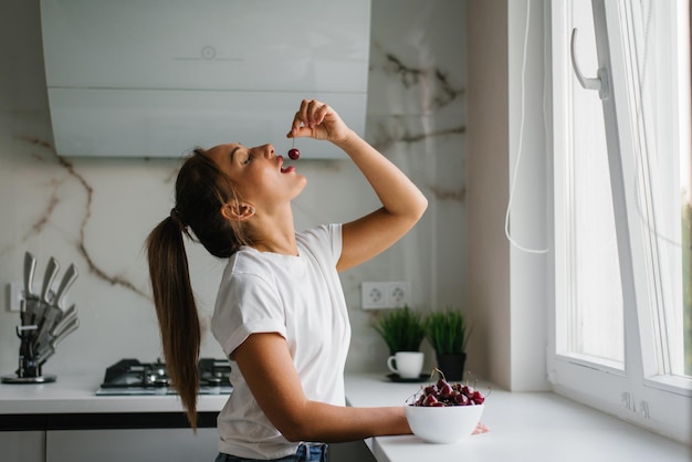 Attractive young woman eats fresh cherries healthy berries sitting at home in the kitchen