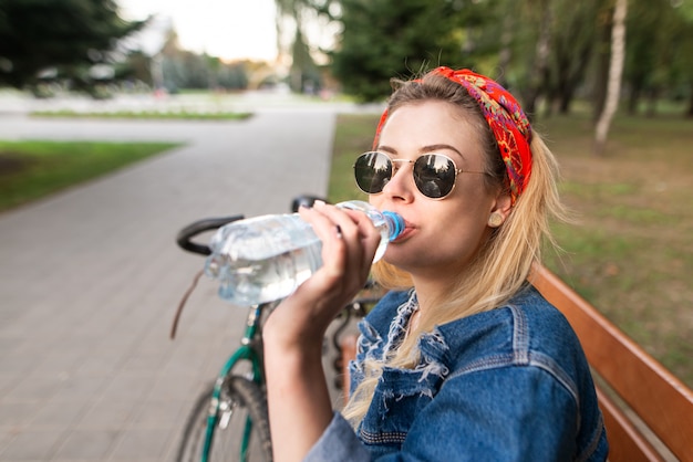 Attractive young woman in glasses sitting on a bench in the park and drinking water from a bottle.