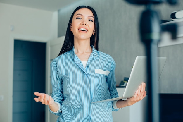 Attractive young woman holding a laptop and smiling while gesturing to the camera for her video blog