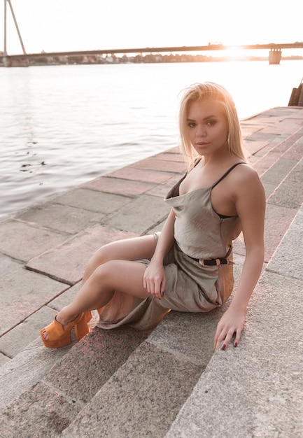 Attractive young woman is sitting on stairs near river at bright sunny day.