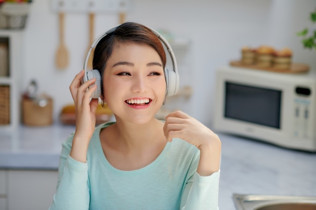 Attractive young woman listening to music with mobile phone and headphones while having breakfast in the kitchen