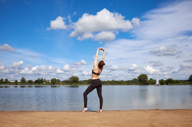 Attractive young woman or model wearing stylish black sportswear practicing yoga and stretcing on the sand near lake. Outdoor activities.