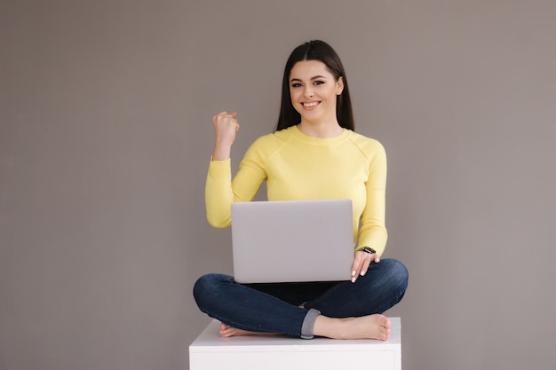 Attractive young woman sitting with laptop on gray background.