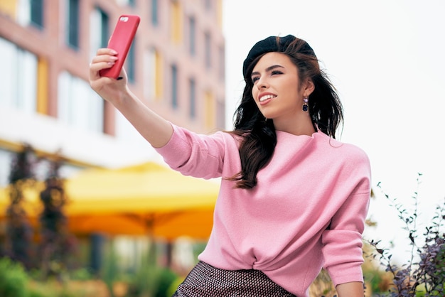 Attractive young woman standing outdoors and taking self portrait on smart phone Brunette female dressed in pink sweater black beret making selfie on the city street