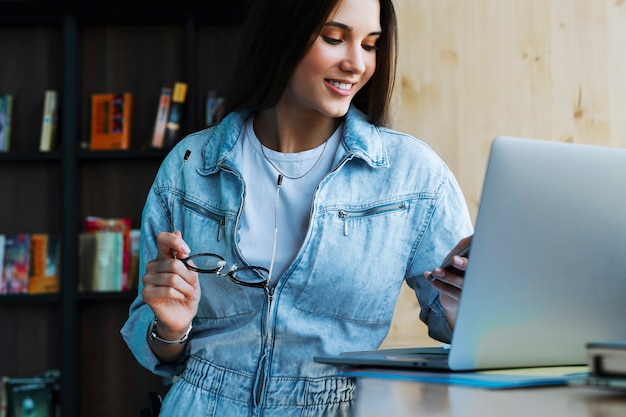 Attractive young woman stands near the window, holds smartphone in her hand, glasses for vision and works on laptop.