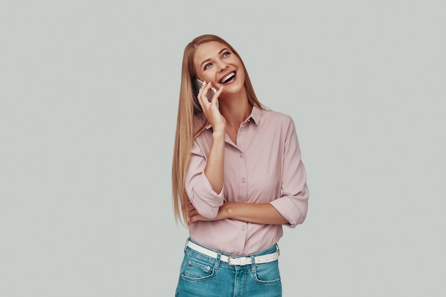 Attractive young woman talking on the smart phone and smiling while standing against grey background