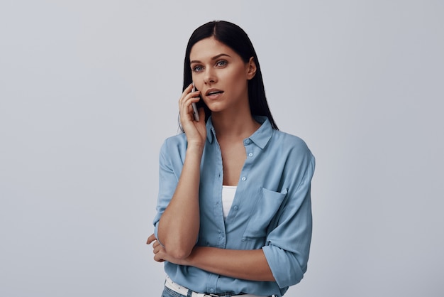 Attractive young woman talking on the smart phone while standing against grey wall