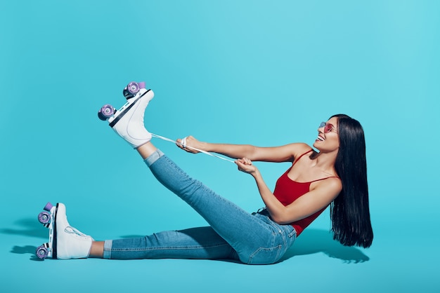 Attractive young woman tying roller skate and smiling while sitting against blue wall