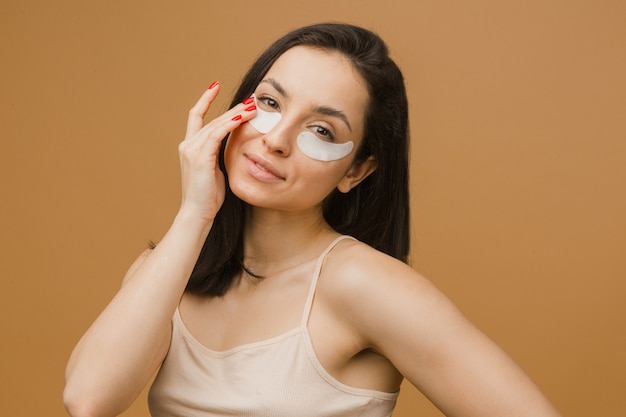 Attractive young woman in underwear with patches on eyes while standing against beige background