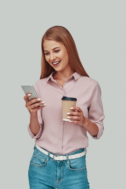 Attractive young woman using smart phone and smiling while standing against grey background