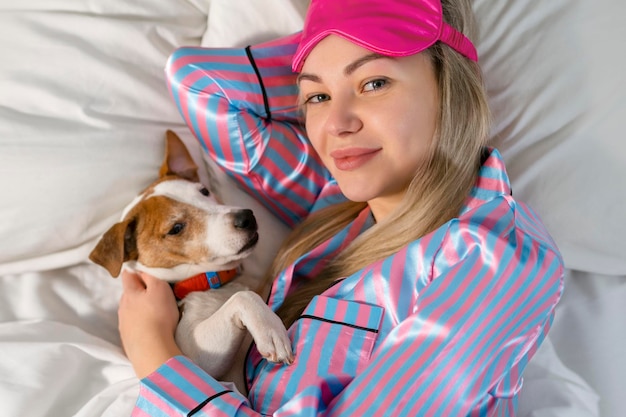 An attractive young woman wearing pajamas is holding a dog while laying on a bed