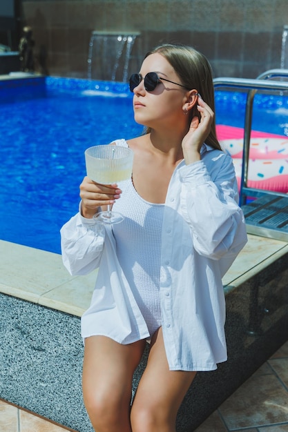 Attractive young woman in a white swimsuit sitting by the pool and drinking a delicious cocktail and sunbathing by the pool during summer vacation