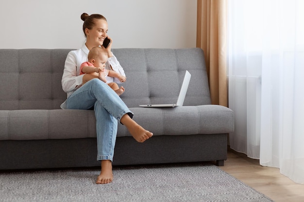 Attractive young woman with bun hairstyle in white t shirt sitting on sofa with baby daughter female freelancer working on laptop and talking with partner on mobile phone