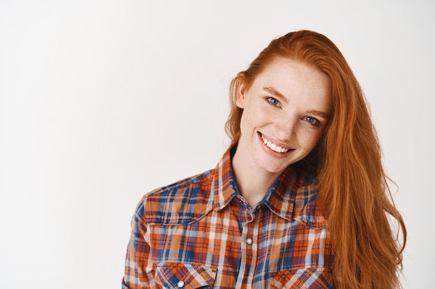 Attractive young woman with ginger hair and freckles, looking at front with perfect smile, standing in shirt against white wall