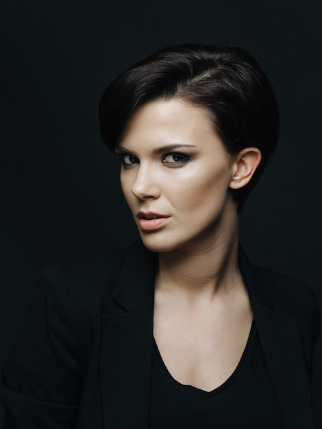 Attractive young woman with light makeup and stylish haircut posing over black wall. Successful lady wearing elegant business clothes.