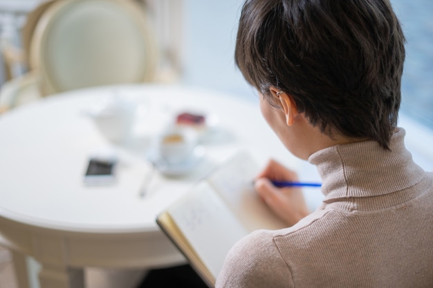 Attractive young woman with pen in hand thinking on plans and writing list to do in notepad enjoying recreation time in stylish cafe