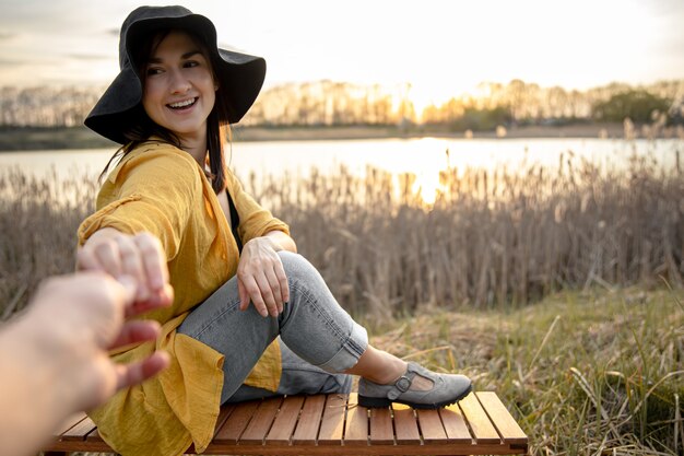 Attractive young woman with a smile on her face holds the hand of the photographer near the lake at sunset