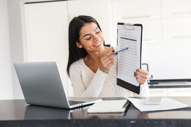 Attractive young woman working with laptop computer and documents while sitting at the kitchen, showing graph