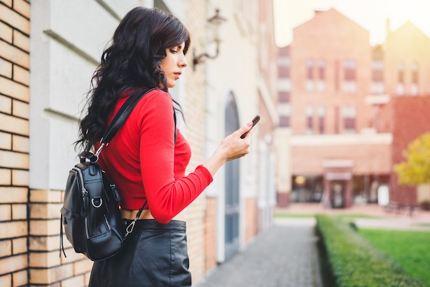 Attrative dark-haired tourist girl walking through the old town street with mobile phone in hands, browsing map application