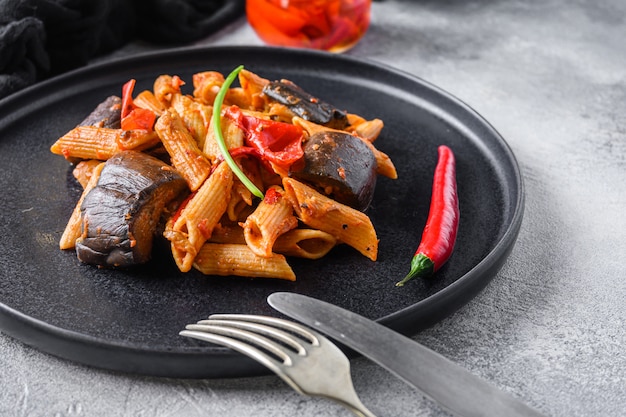 Aubergine penne eggplant pasta, pepper tomatoe sauce, on black plate overgrey concrete background  served side view.