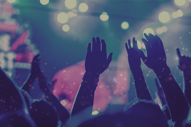 Audience with hands raised at a music festival and lights streaming down from above the stage.