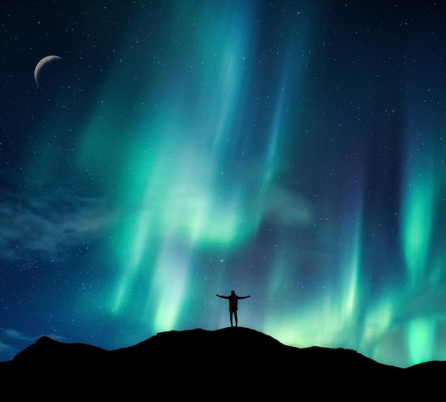 Aurora borealis glowing over silhouette hiker standing on the mountain in the night sky