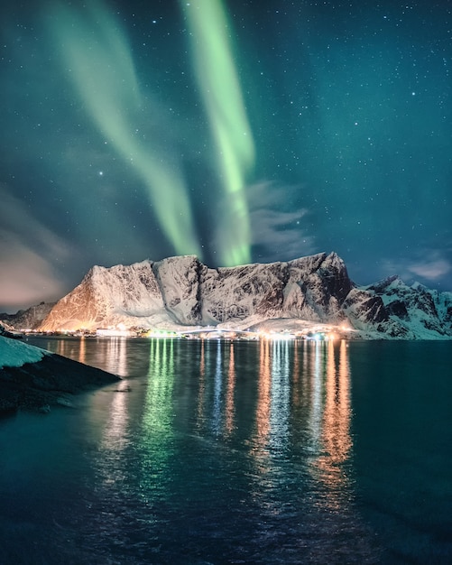 Aurora Borealis, Northern Lights over snowy mountain with glowing village in Hamnoy at Lofoten Islands, Norway