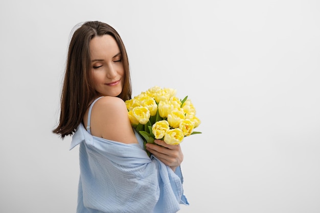 Authentic portrait of caucasian woman with closed eyes in blue clothes with bouquet of yellow tulip flowers stands sideways on white background