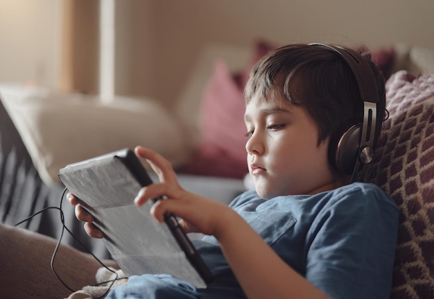 Authentic portrait Kid sitting on sofa watching cartoons on tabletYong boy playing game on touch pad Child lying on couch wearing headphoes listening to music or relaxing on his own in living room